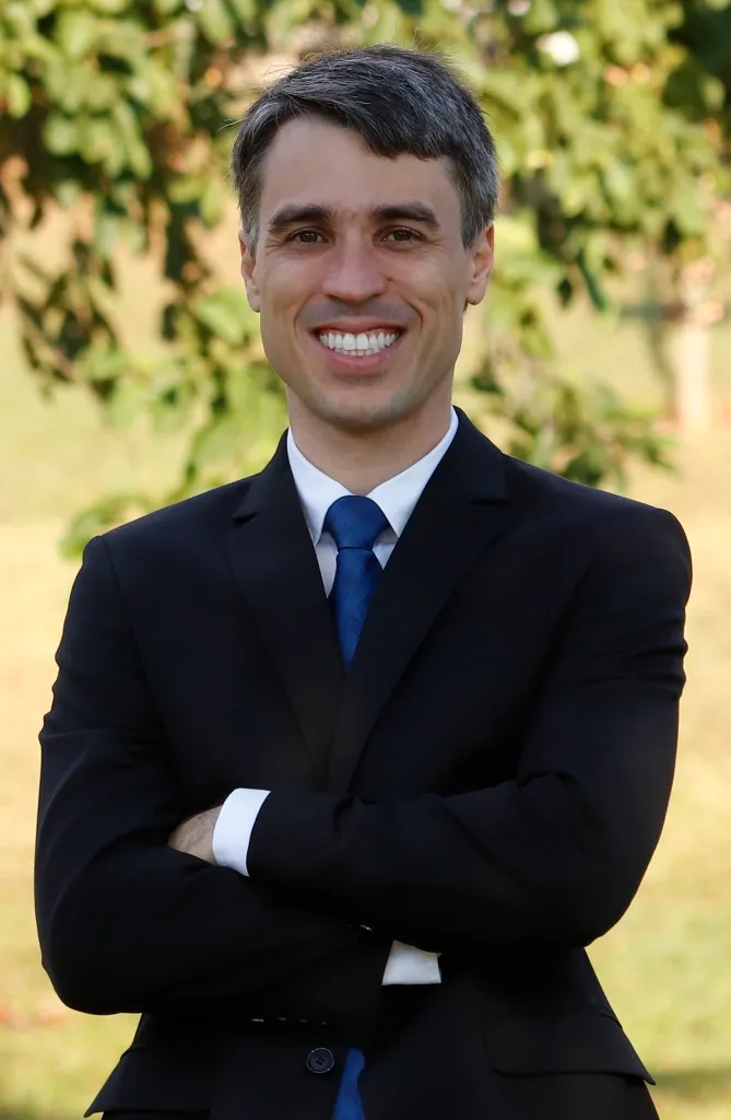 Retrato profissional de Pedro Rafael de Moura Meireles, advogado fundador, terno escuro azul-marinho, sorriso confiante, braços cruzados, fundo natural desfocado com folhagem verde, iluminação natural suave, fotografia corporativa realista